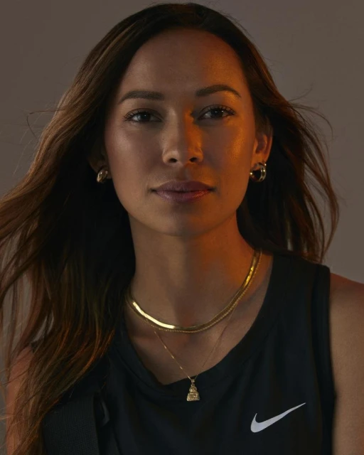 Portrait of an adult woman with long brown hair, wearing a black tank top, gold necklaces, and hoop earrings, against a grey backdrop.