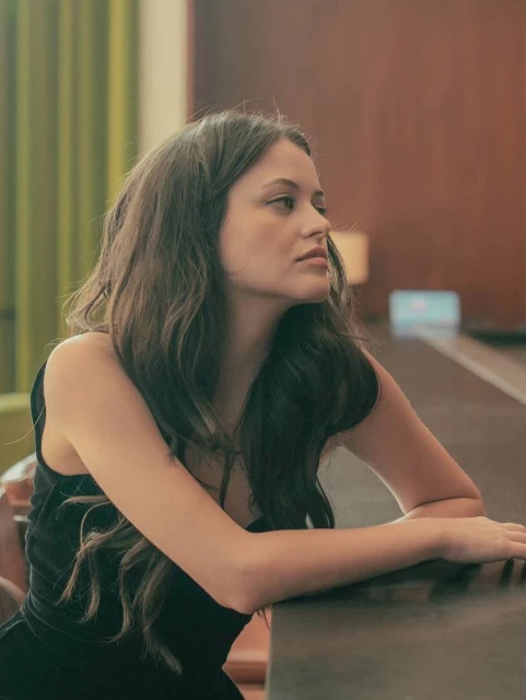Portrait of a young adult woman in a black dress looking off camera, with wavy brunette hair and a neutral expression, indoor.
