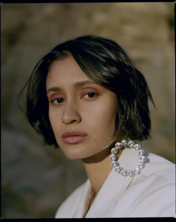 Close-up portrait of a woman with dark hair wearing crystal hoop earrings and a white jacket against a natural rocky background.