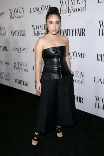 A woman in a black strapless leather top and wide-legged pants poses at an event with sponsor logos visible in the background.