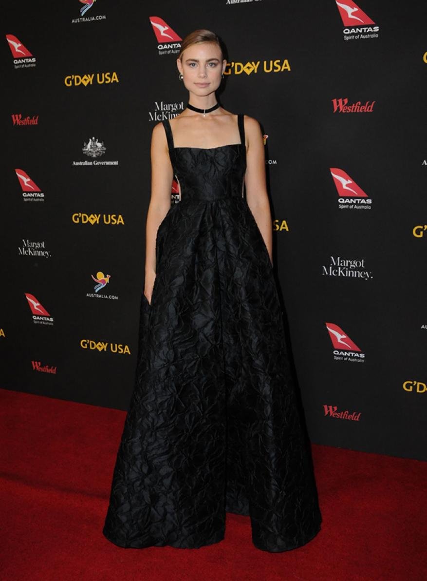 Woman in black gown on red carpet at G'Day USA event. Evening light, formal wear, celebrity appearance, black dress, red carpet, event backdrop.