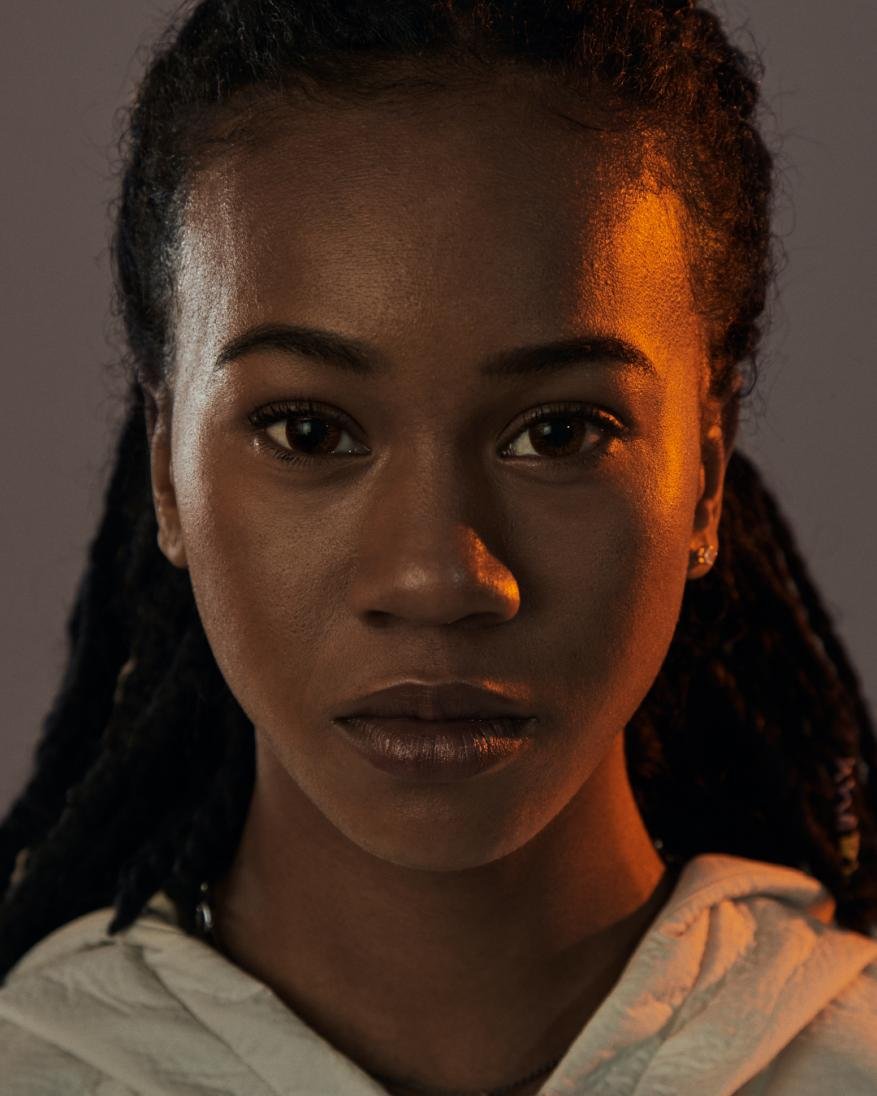 Close-up portrait of an adult Black woman with dreadlocks, wearing a white hoodie, against a gradient grey background with orange side lighting.
