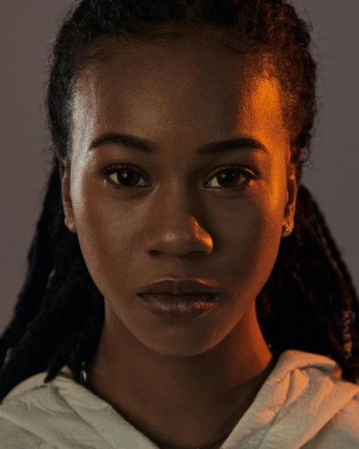 Close-up portrait of an adult Black woman with dreadlocks, wearing a white hoodie, against a gradient grey background with orange side lighting.
