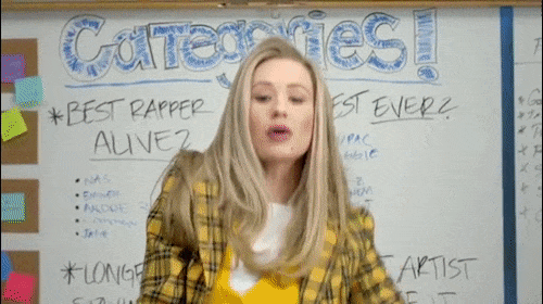 Woman in a yellow plaid blazer poses in front of a whiteboard with hand-written categories and lists in a classroom setting.