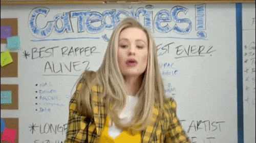 Woman in a yellow plaid blazer poses in front of a whiteboard with hand-written categories and lists in a classroom setting.