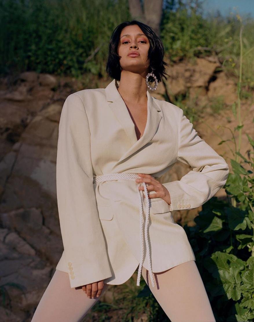 A woman poses outdoors wearing a cream-colored blazer, white tights, and oversized pearl earrings, set against a backdrop of rock and foliage.