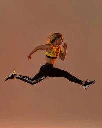 Female athlete captured mid-air in a dynamic running pose wearing a yellow sports bra and black leggings against a neutral studio backdrop