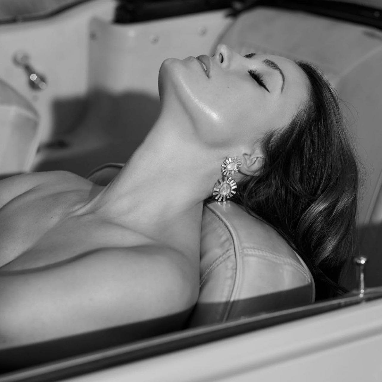 Monochrome portrait of an adult woman with elegant earrings, lying back in a classic car with eyes closed, and soft lighting.