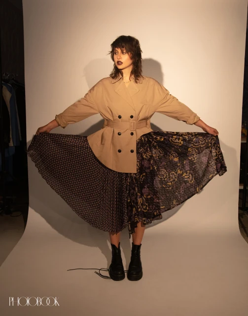 Full-length studio shot of a woman wearing a tan double-breasted jacket, asymmetrical floral print dress, and black boots against white backdrop.