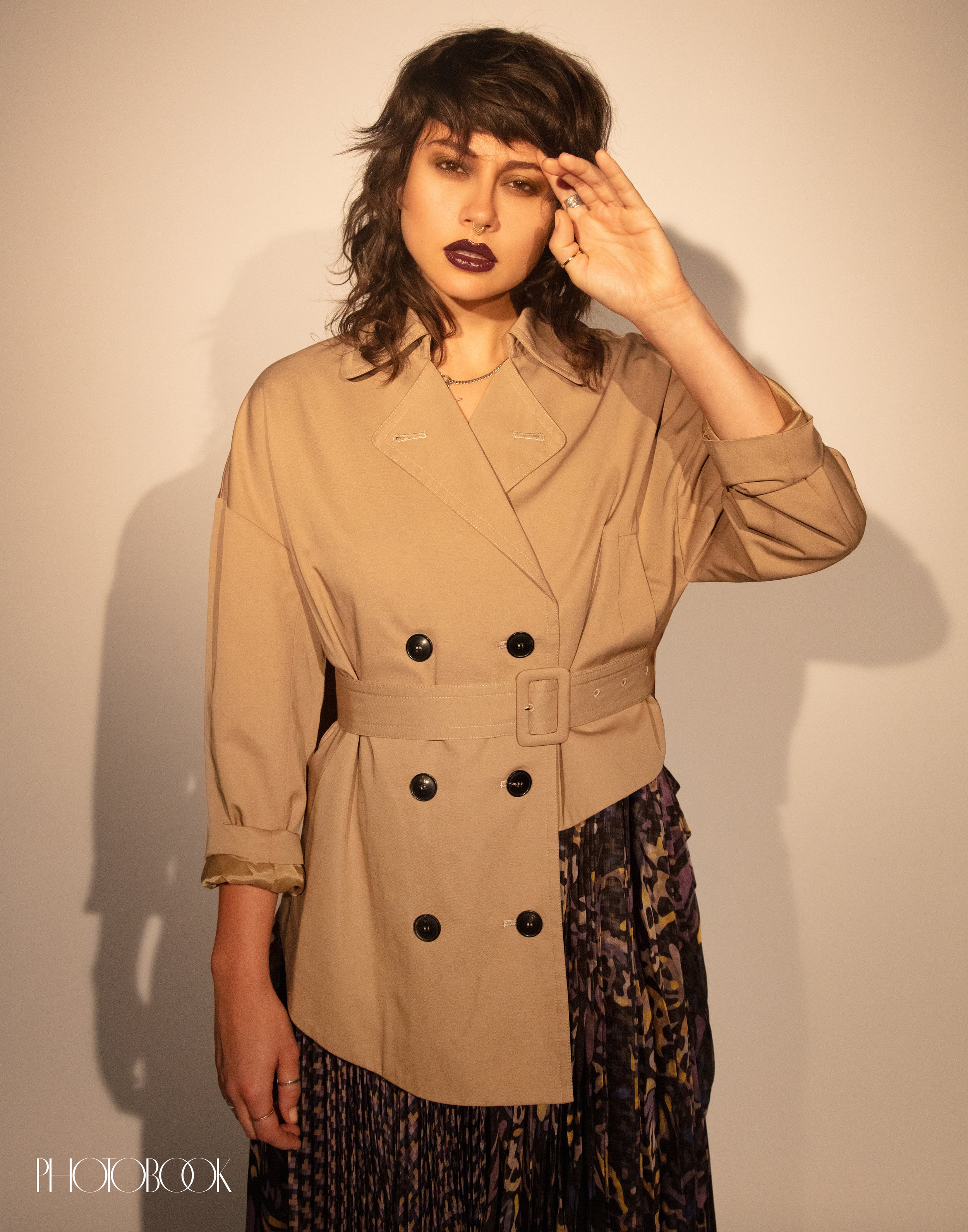 Adult woman with dark lipstick and a beige belted jacket poses in front of a neutral backdrop. Fashion editorial portrait.