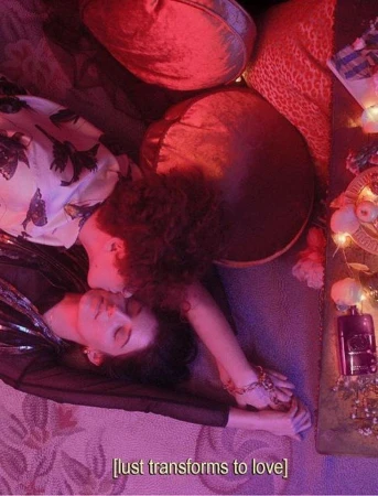 Two women are lying on a floral patterned surface with a red hue, close together, with gold accessories and a perfume bottle visible.