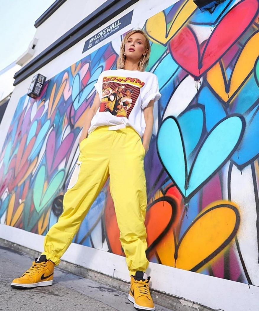 Young woman in yellow pants and a white t-shirt standing in front of a colorful graffiti wall in an urban outdoor setting.