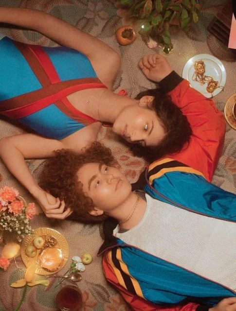 Two people lie on a patterned surface among teacups, fruit, and flowers, in a warm and artistic indoor setting with a top-down view.