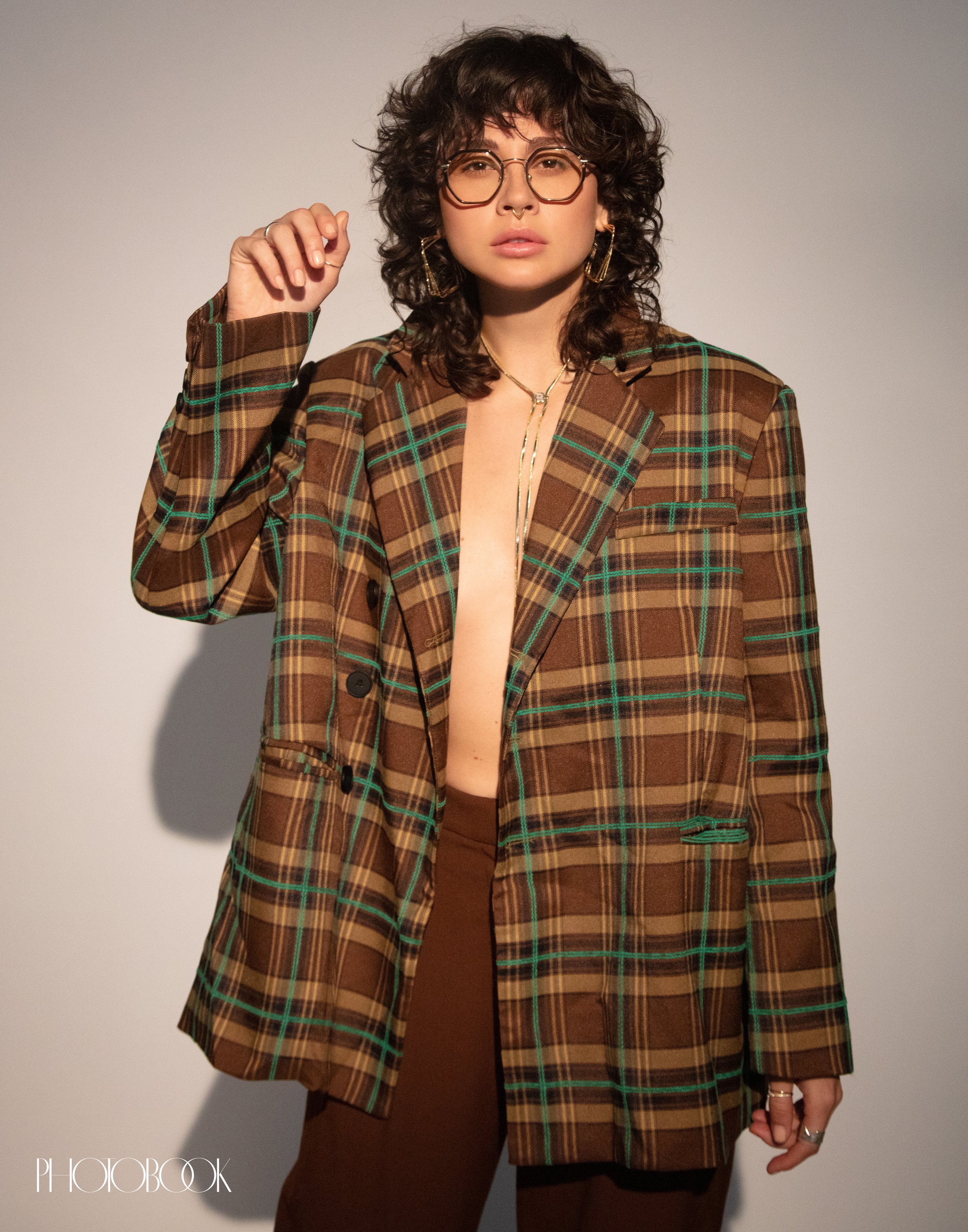 A woman with curly hair wearing a brown plaid blazer and brown pants poses in front of a white wall in a studio setting.