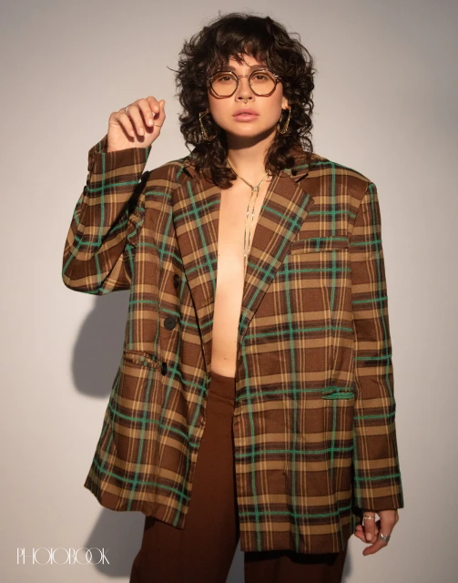 A woman with curly hair wearing a brown plaid blazer and brown pants poses in front of a white wall in a studio setting.