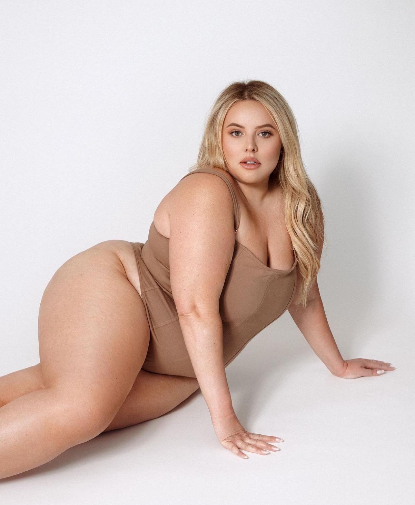 Blonde woman poses in a studio wearing a brown one-piece swimsuit. She is lying on her side with her hands on the white floor.