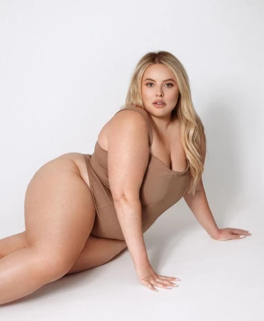 Plus size blonde model wearing a brown bodysuit posing against a clean white seamless studio background.