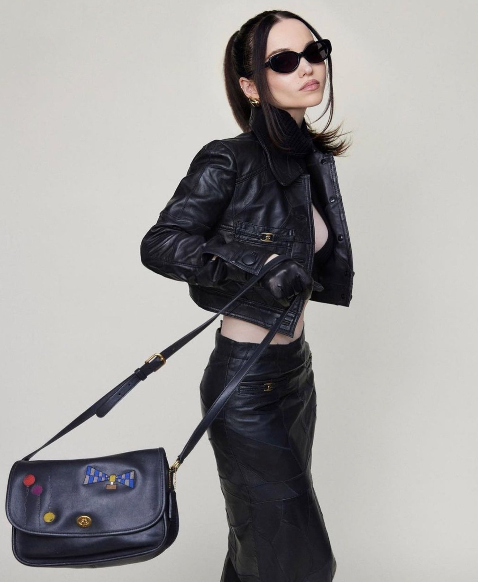 Fashionable woman in black leather jacket, skirt, and gloves poses with a decorated black shoulder bag in a studio setting.