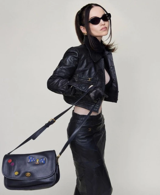 Fashionable woman in black leather jacket, skirt, and gloves poses with a decorated black shoulder bag in a studio setting.