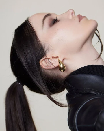 Profile portrait of a woman with dark hair in a ponytail wearing a gold teardrop earring and a black leather jacket against a light backdrop.