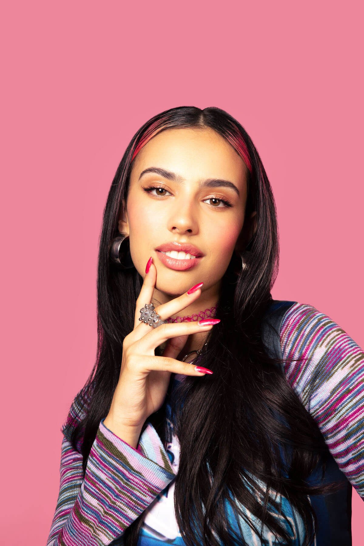 Portrait of a young woman with dark hair and hoop earrings, wearing a patterned jacket, posing against a solid pink background.