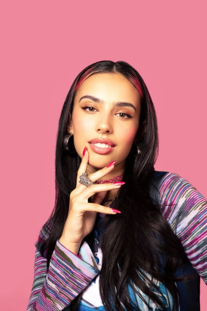 Portrait of a young woman with dark hair and hoop earrings, wearing a patterned jacket, posing against a solid pink background.