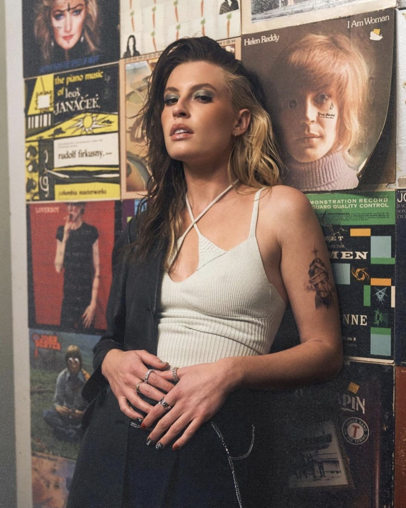 A woman with two-toned hair poses in front of vintage record covers in a medium shot. She wears a black jacket and a white top.