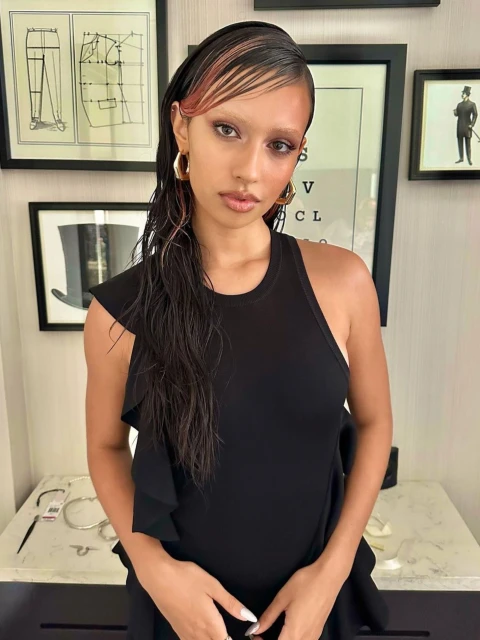 Portrait of a woman with dyed eyebrows and wet hair poses indoors wearing a black dress, with framed artwork visible in the background.