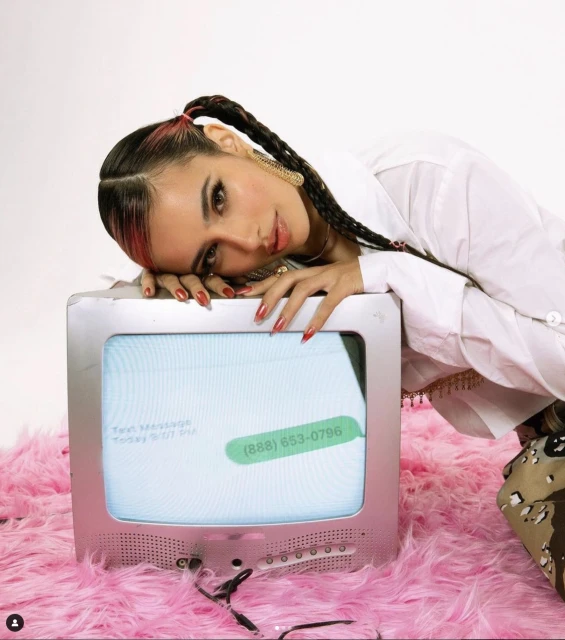 A woman with braided hair rests her head on a retro TV displaying a phone number against a white background and a pink fluffy rug.