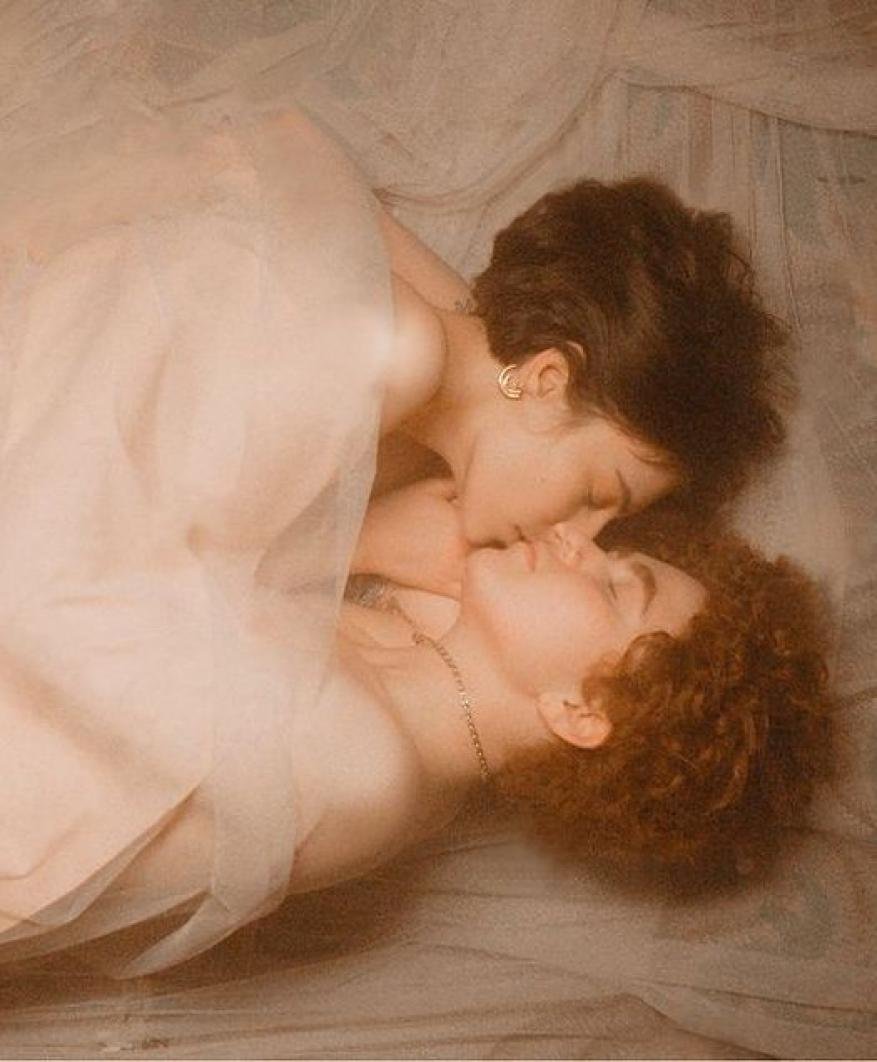 Two women lie intimately together, wrapped in sheer fabric, in soft, diffused lighting. The focus is on their close connection and embrace.
