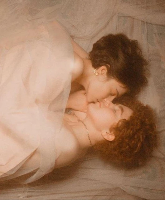 Two women lie intimately together, wrapped in sheer fabric, in soft, diffused lighting. The focus is on their close connection and embrace.