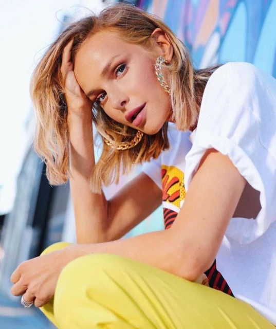 Blonde woman poses in yellow pants and white t-shirt, leaning against a colorful graffiti wall in a medium shot with chain earrings.