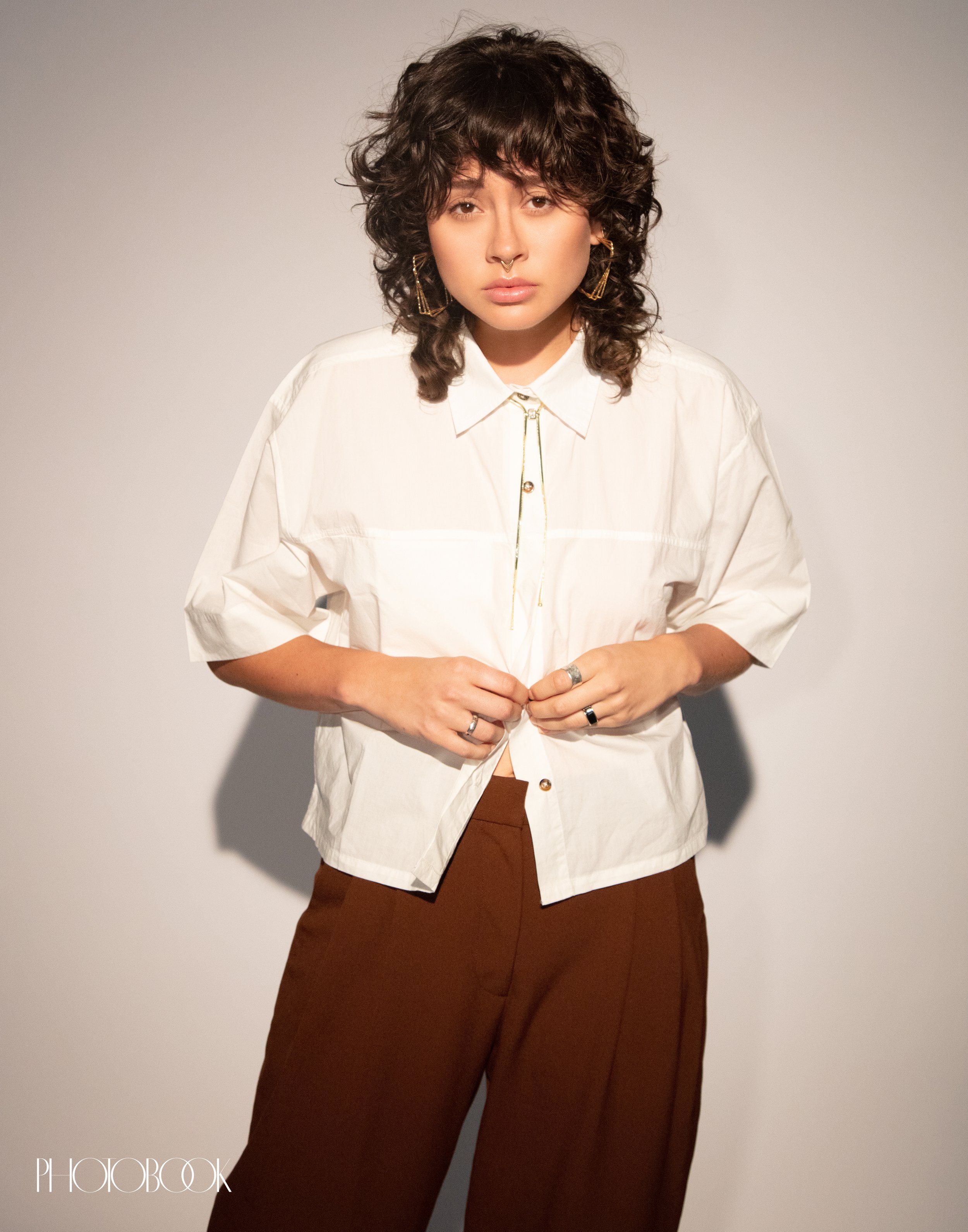 Young woman in a white button-up shirt and brown pants poses in front of a grey background, accessorized with gold earrings and rings.