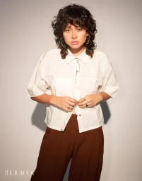 Portrait of a person with dark curly hair wearing a white short-sleeve button-down shirt and brown trousers against a plain white background.