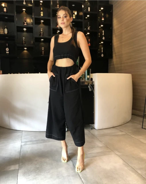 Young woman in stylish black crop-top jumpsuit poses indoors against a modern bar backdrop, showcasing a contemporary fashion look.