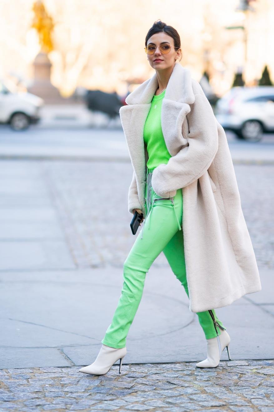 Woman walks on a city street in a neon-green outfit, white coat, and matching boots. A blurry city background with vehicles is present.