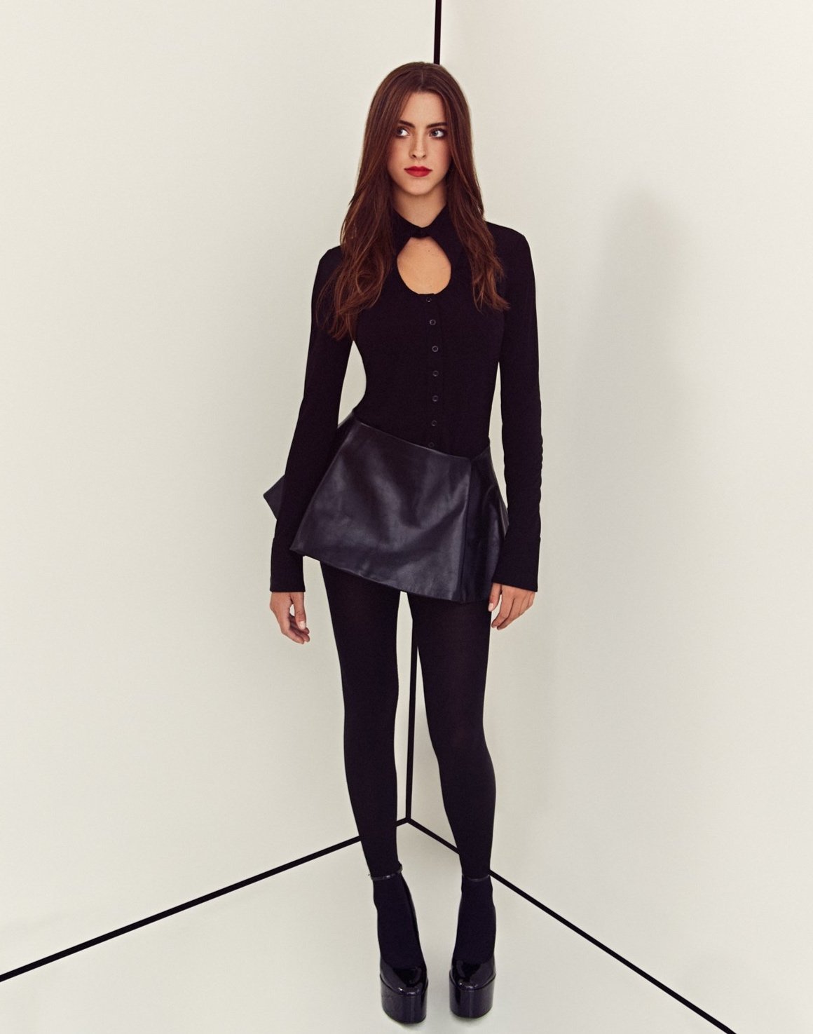 A woman in a black top and leggings stands in the corner of a studio with bright lighting, showcasing fashion-forward attire and platform shoes.