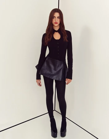 A woman in a black long-sleeve top, skirt, tights and platform heels standing in a white corner.