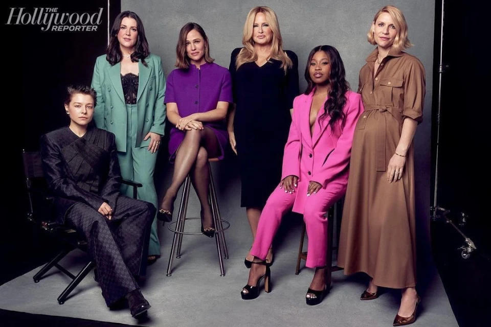 Six women, dressed in fashionable suits and dresses, pose indoors for a portrait on a grey backdrop with visible studio lighting equipment.