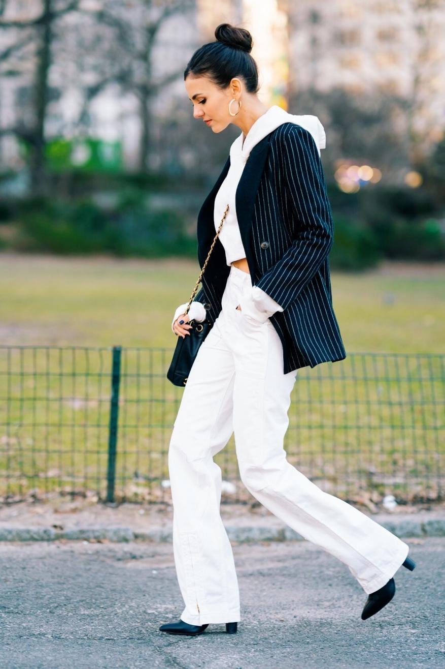A woman walks outdoors wearing a pinstripe blazer, white crop top, and white pants, accessorized with gold hoop earrings and a shoulder bag.