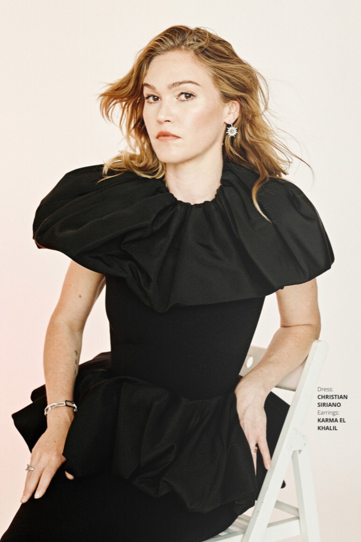 Woman with shoulder-length blonde hair poses in a black dress with a large collar while sitting on a white chair against a white backdrop.