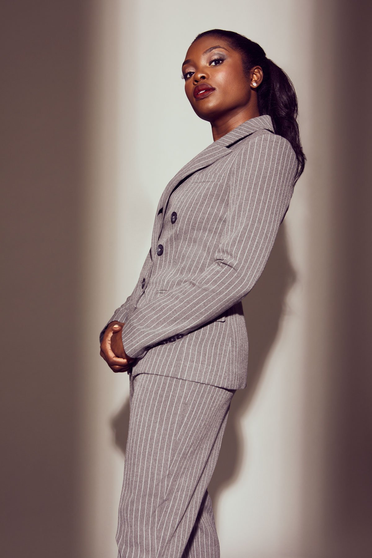 Portrait of a young Black woman in a stylish grey pinstripe pantsuit, posing indoors with a confident expression and soft lighting.