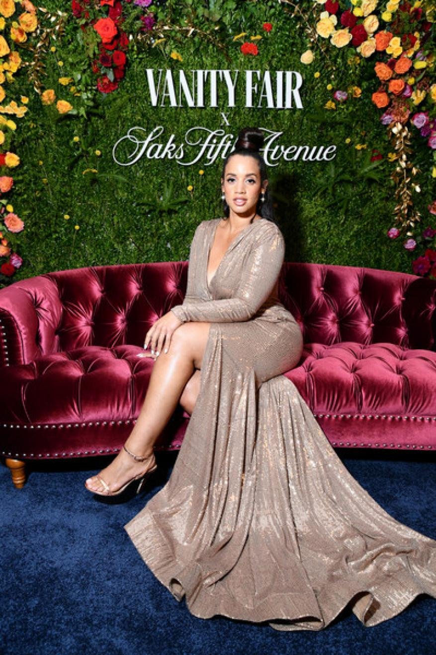 Woman poses on a velvet tufted couch at a Vanity Fair and Saks Fifth Avenue event with a floral backdrop in a long sequin gown.