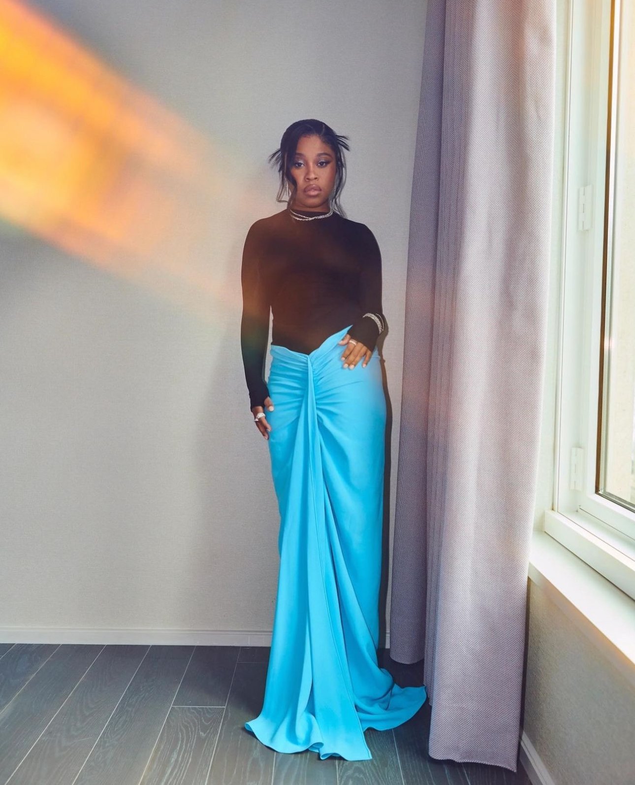 A stylish woman in a long-sleeved black top and draped bright blue skirt poses against a neutral wall with a window light effect.