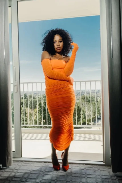 Woman in an orange dress and matching gloves stands framed by an open doorway to a balcony with a scenic tree view on a sunny day.