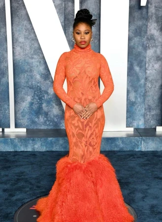 An elegant African-American woman poses in a long-sleeved orange gown with a fur-like train against a mottled blue and white backdrop.
