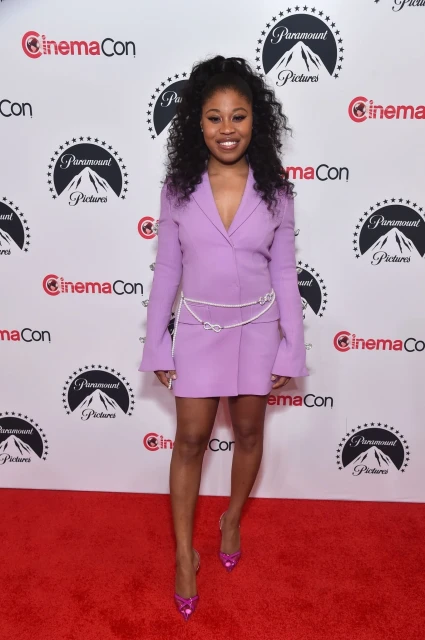 A smiling Black woman in a lilac blazer dress poses on a red carpet in front of a backdrop with Paramount Pictures logos at CinemaCon.