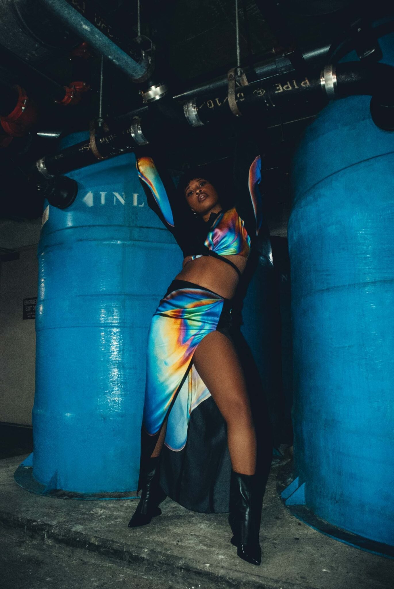 A woman in a multicolored outfit poses in a basement-like area with large blue tanks and overhead pipes, creating a fashion-forward image.