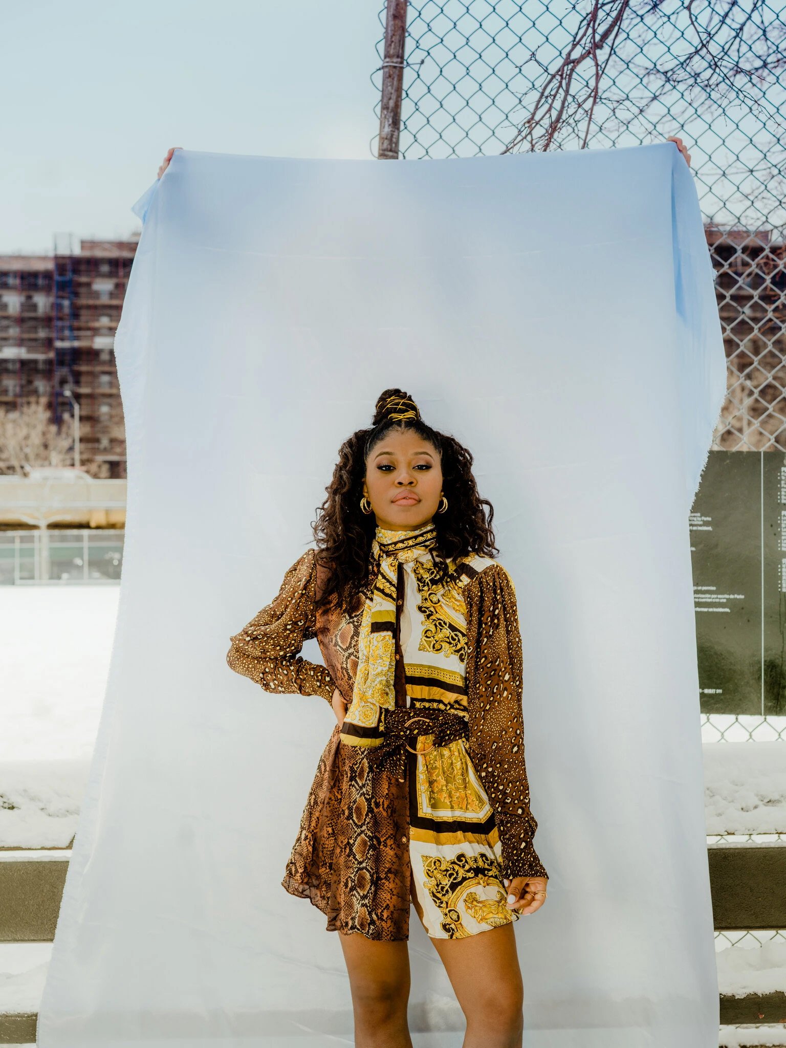 A stylish Black woman poses outdoors in a patterned dress and scarf against a white sheet, with a fence and building in the background.