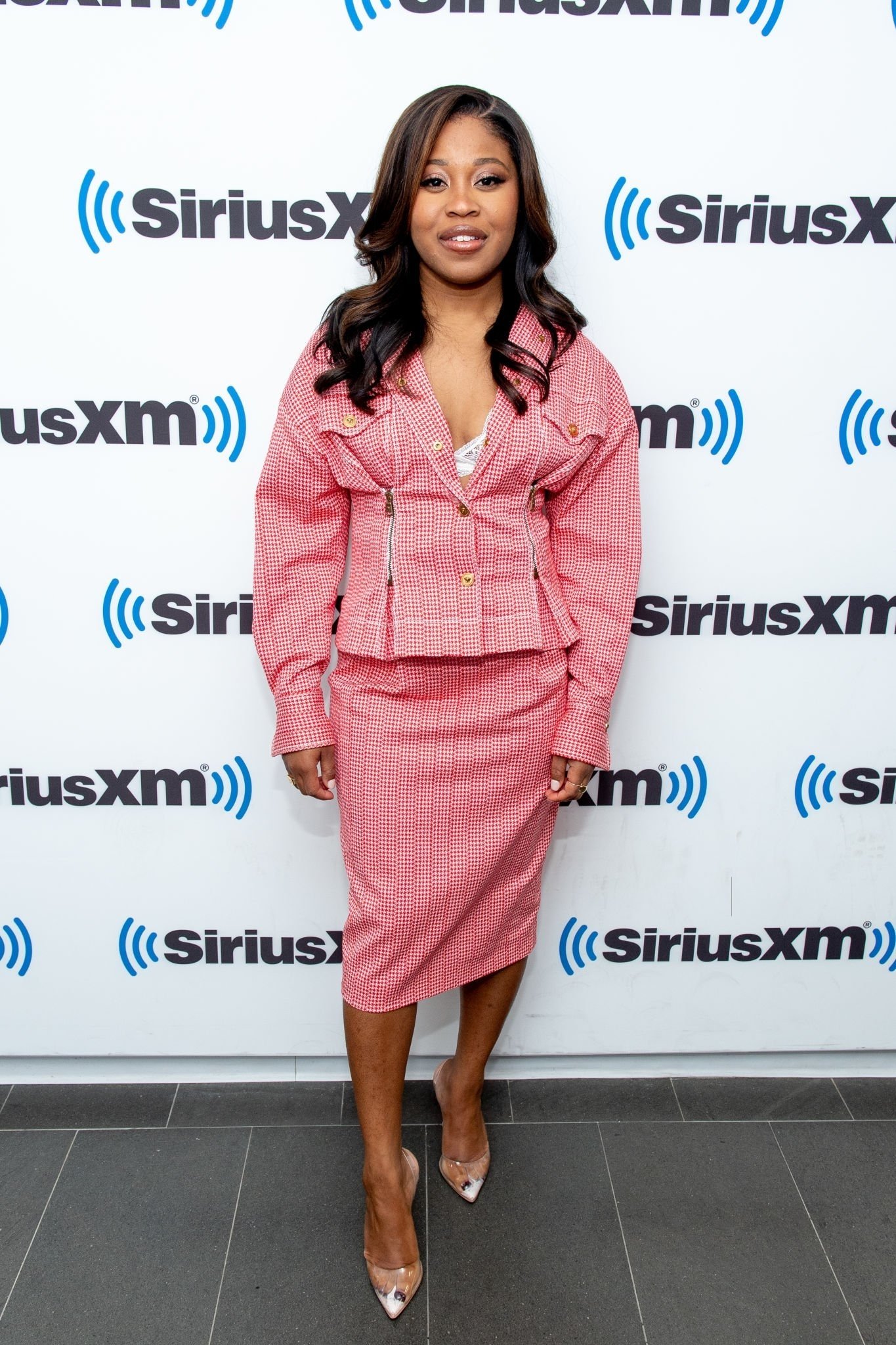 Attractive African American woman in pink skirt suit smiles, posing in front of SiriusXM logo, wearing clear pointed toe heels.